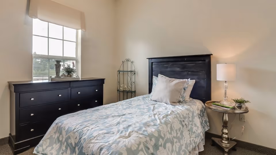 A neatly made single bed with a black wooden headboard and light blue floral bedding in a small bedroom. Next to the bed is a round silver side table with a lamp, a small plant, and some magazines. Against the wall under a window with white blinds is a black dresser with multiple drawers. A decorative metal shelf stands in the corner between the dresser and the bed.