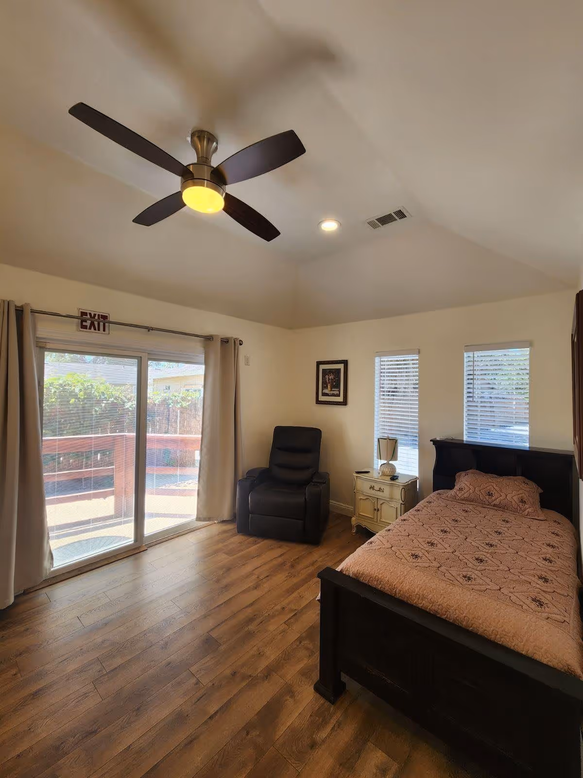 A bedroom with wooden flooring, a single bed with a patterned bedspread, a black recliner chair, a small white nightstand with a lamp, two narrow windows with blinds, and a sliding glass door with curtains leading to an outdoor area. A ceiling fan with a light is mounted on the ceiling.