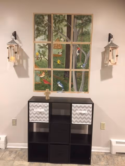 A small black shelving unit with two gray and white chevron-patterned storage bins is placed against a beige wall. Above the shelving unit is a decorative wall art piece resembling a window with nine panes, depicting a tree with various colorful birds and a birdhouse. On either side of the wall art, there are two lantern-style wall sconces with candles inside. The floor is tiled in a light color.