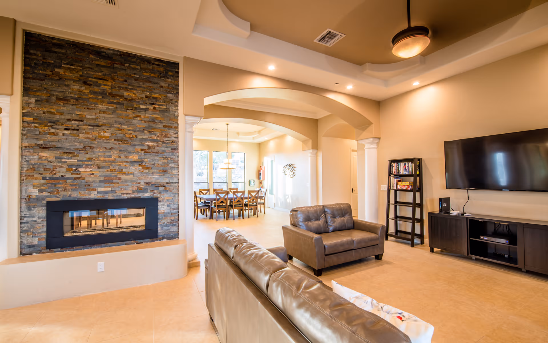 A spacious living room area featuring a modern stone fireplace on the left, two brown leather sofas, a ceiling fan with light, a flat-screen TV mounted on the wall, and a black shelving unit with books and games. In the background, there is a dining area with a wooden table and chairs under a hanging light fixture, illuminated by natural light from large windows.