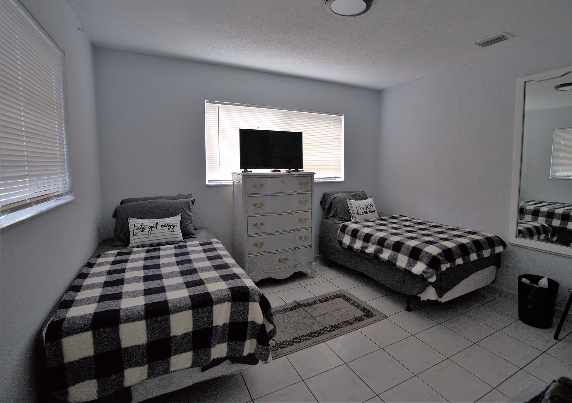 A small bedroom with two single beds, each covered with a black and white checkered blanket and a decorative pillow. Between the beds is a white chest of drawers with a flat-screen TV on top. The room has white tiled floors, white walls, a large window with blinds behind the chest of drawers, and a full-length mirror on the right wall. A small rug is placed on the floor between the beds.