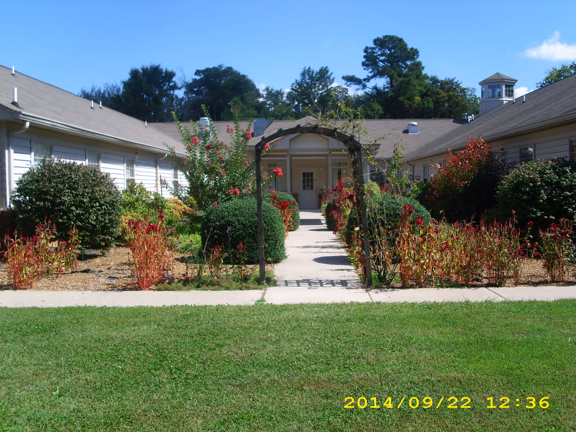 View of a garden courtyard with a concrete pathway leading through an archway covered with climbing plants, surrounded by bushes and red flowers, flanked by two single-story buildings under a clear blue sky.