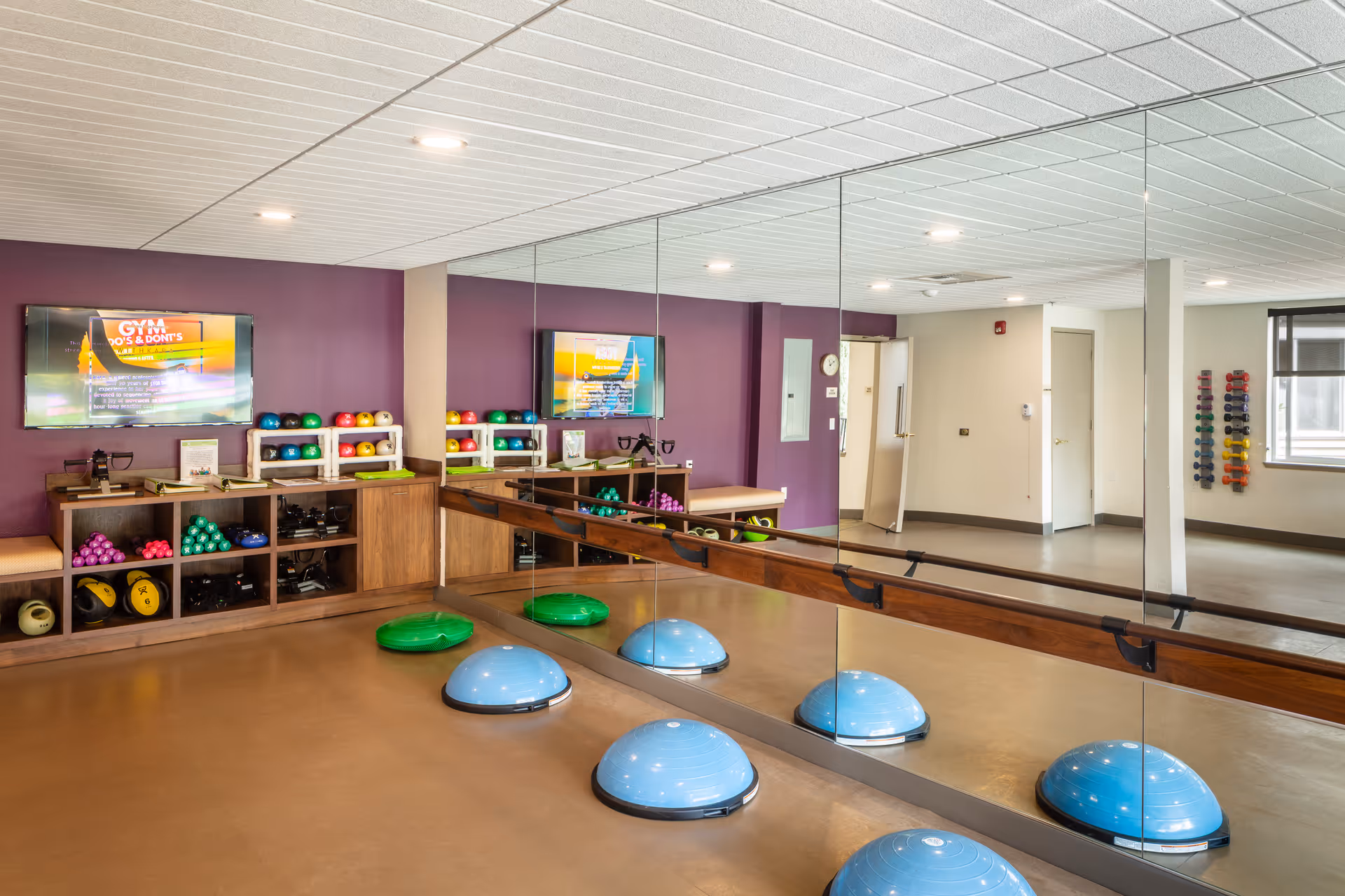 Bright fitness room with mirrored wall, balance domes on the floor, colorful dumbbells and a TV mounted on a purple wall.