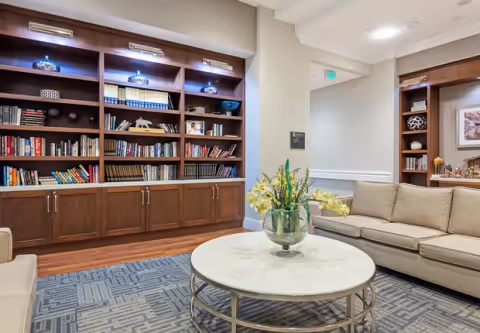 A cozy living room area in Tribute at The Glen featuring a large wooden bookshelf filled with books and decorative items, a beige sofa, a round white coffee table with a glass vase holding yellow flowers, and a patterned gray rug on a wooden floor.