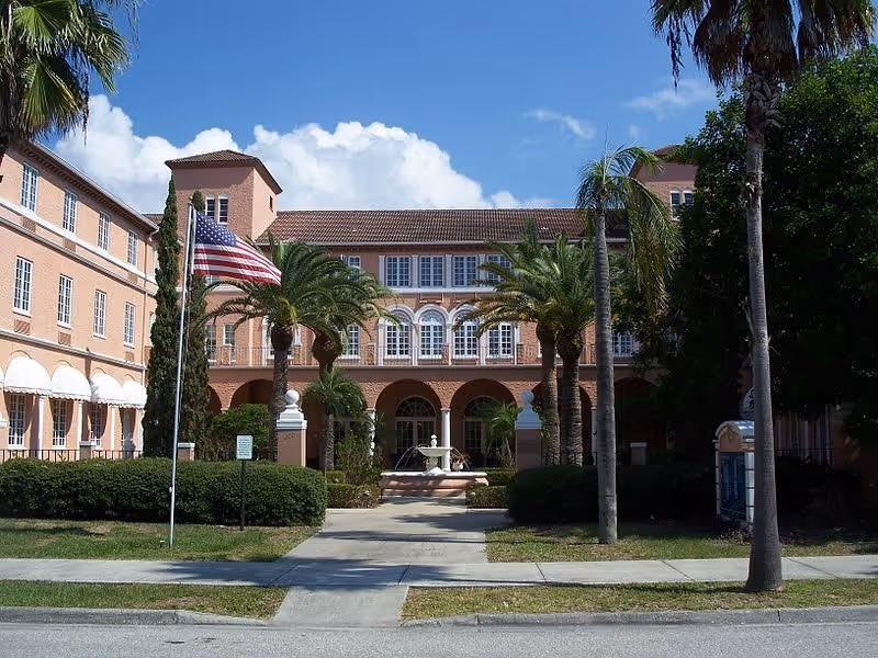 Exterior view of The Pavillion of Downtown Venice, a large peach-colored building with multiple windows and arches. The entrance is framed by palm trees and an American flag on a flagpole. There is a fountain in front of the entrance and a clear blue sky with some clouds above.
