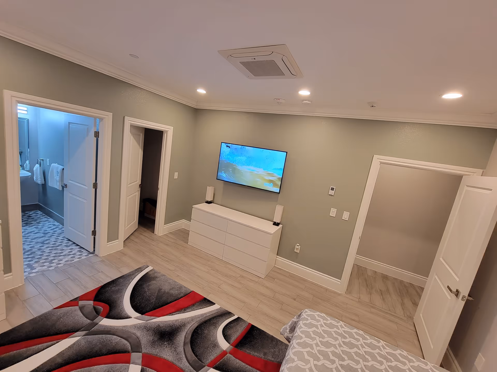 Interior view of a bedroom in an assisted living facility with a patterned rug on the floor, a bed partially visible with a gray and white patterned bedspread, a wall-mounted flat screen TV above a white dresser with two speakers, and two open doorways leading to a bathroom and a closet.