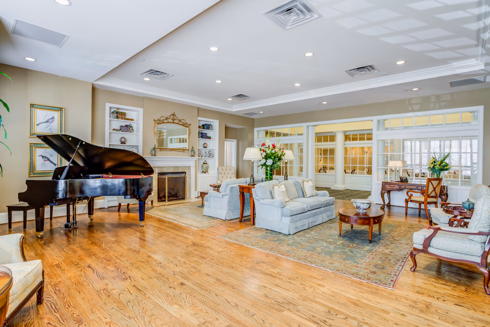 A spacious and elegant living room area with polished hardwood floors, a grand piano, a fireplace with a decorative mirror above it, built-in bookshelves, and comfortable seating including sofas and armchairs arranged around a coffee table. The room is well-lit with recessed ceiling lights and table lamps, and large windows with white trim provide a view into an adjacent room.