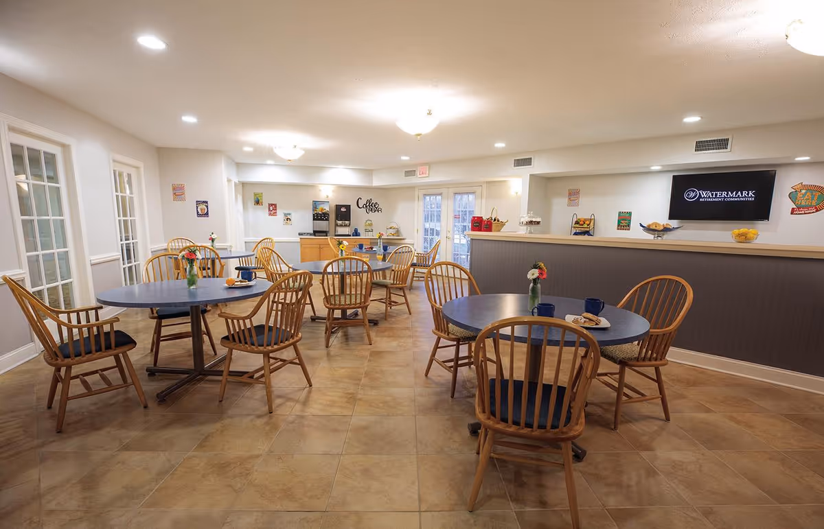 A dining area with several round tables and wooden chairs arranged neatly. Each table has a small vase with flowers and some cups and plates. In the background, there is a coffee bar with a coffee machine and various items on the counter. The room has beige tiled flooring, light-colored walls, and ceiling lights providing bright illumination.
