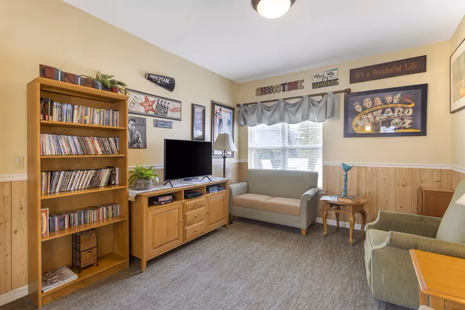A cozy living room with a wooden bookshelf filled with DVDs and CDs, a wooden TV stand with a flat-screen TV, a small sofa, an armchair, and a side table with a lamp. The walls are decorated with movie-themed signs and posters, and a window with a valance lets in natural light.