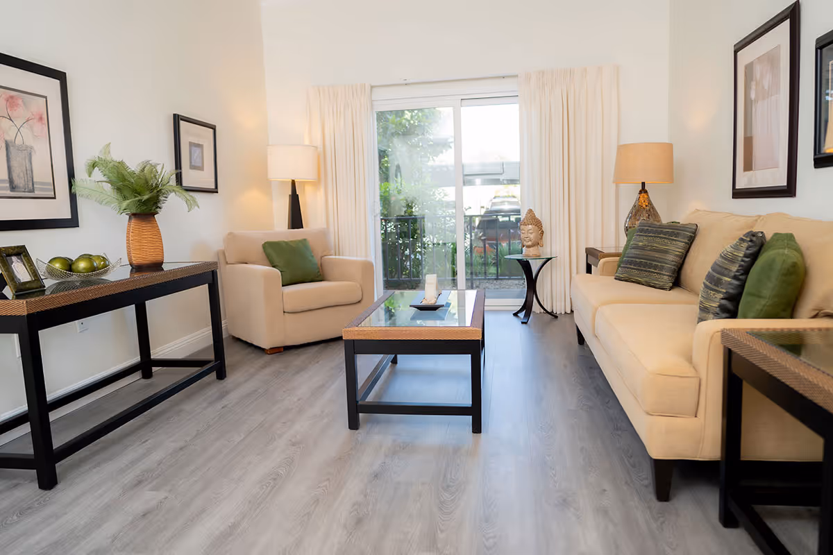 Bright, modern living room with a beige sofa and armchair, coffee table, side tables, lamps, and a sliding glass door to a small balcony.