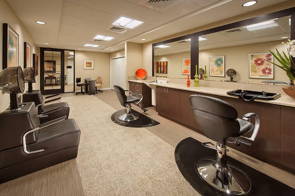 Interior view of a salon area in a senior living facility with black salon chairs, hair dryers, a long counter with a large mirror, decorative vases, and framed floral artwork on the walls. The room has beige walls, a patterned carpet, and a door leading to another room.