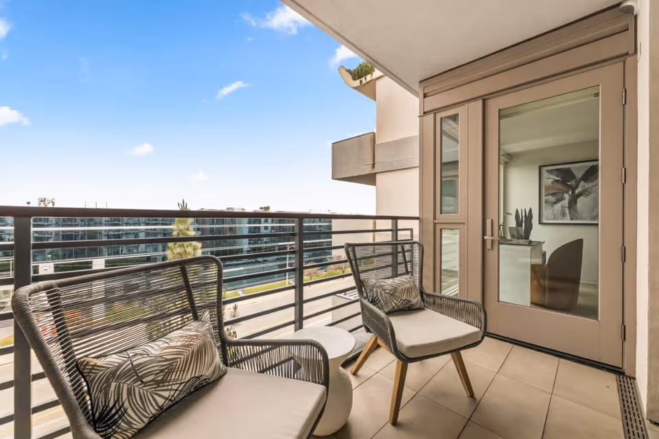 Balcony with two wicker chairs and a small table overlooking a modern building and street, with a glass door into the apartment.