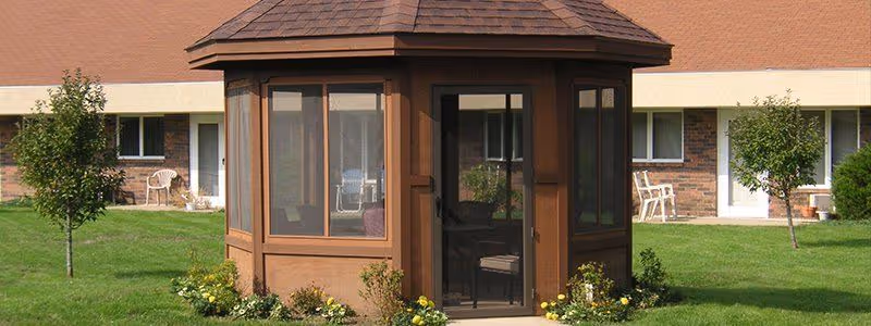 A small brown screened gazebo sits on a grassy courtyard in front of a low brick senior living building with patio chairs and small trees.