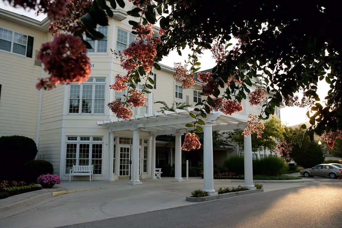 Exterior view of a senior living facility entrance with white columns supporting a pergola. Pink flowers hang from a tree in the foreground, and there are benches and landscaping around the entrance. A car is parked on the right side of the driveway.