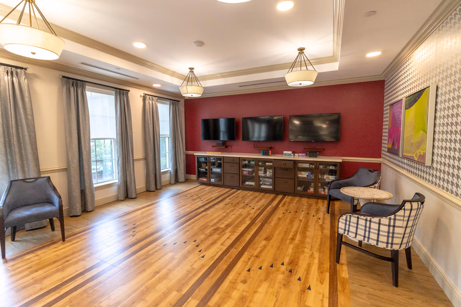 A spacious common room with wooden flooring, three wall-mounted flat-screen TVs on a red accent wall, and a long cabinet below holding board games. The room has three large windows with gray curtains, modern ceiling lights, and several chairs including a plaid armchair and a small round table. The opposite wall features patterned wallpaper and colorful artwork.