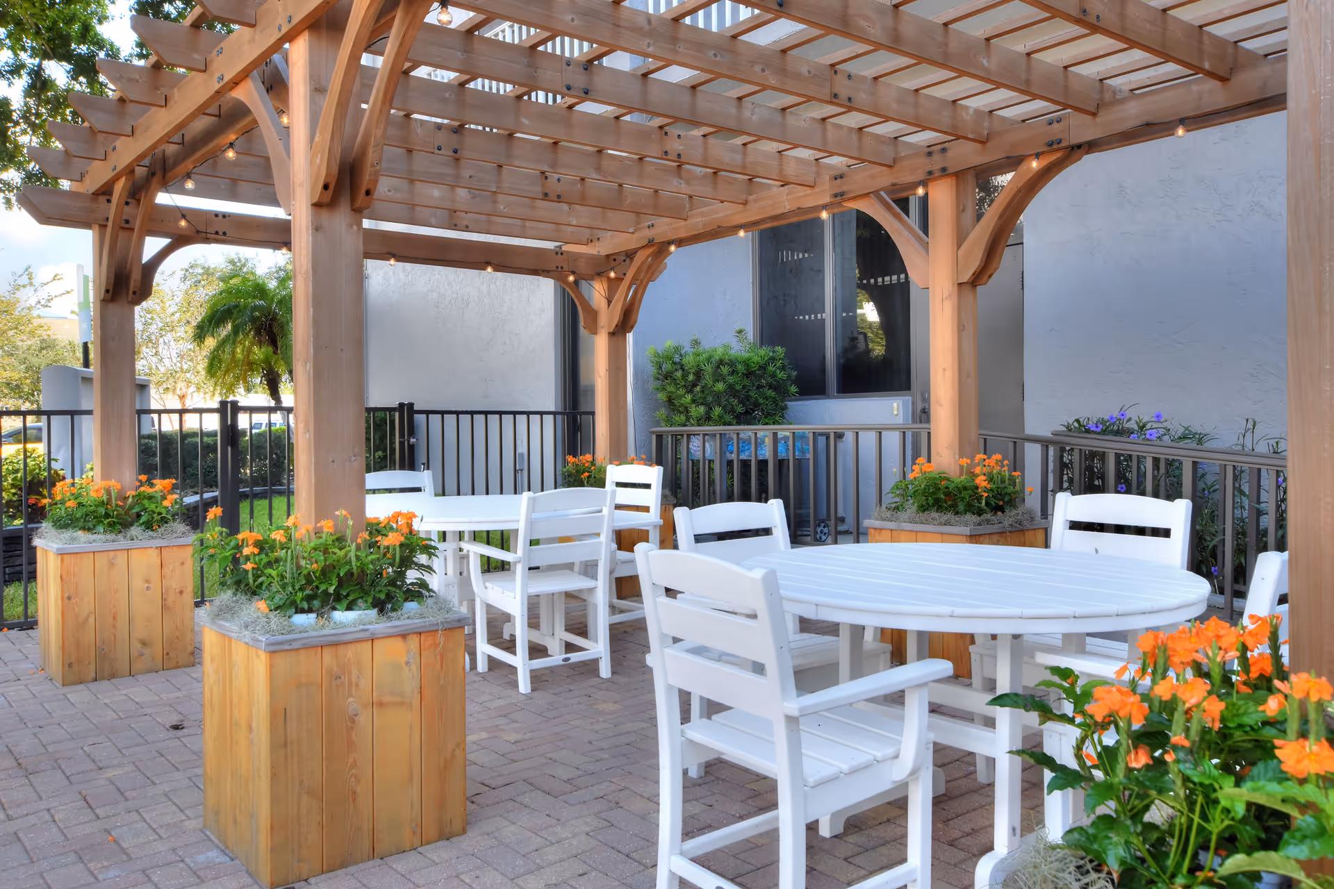 Outdoor patio area with a wooden pergola overhead, white round and square tables surrounded by white chairs, and wooden planter boxes filled with orange flowers. The area is paved with bricks and enclosed by a black metal fence, with some greenery and a building wall in the background.