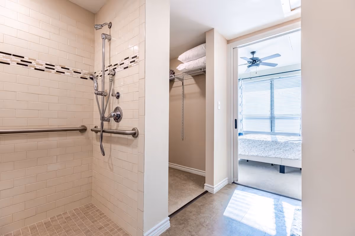 A tiled walk-in shower with grab bars and a handheld showerhead is visible on the left. To the right, there is an open doorway leading to a bedroom with a bed, ceiling fan, and large window with blinds. Between the shower and bedroom is a small closet with shelves holding folded towels.