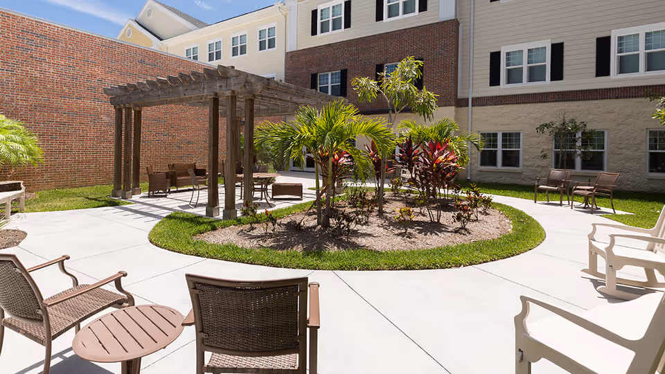Sunny courtyard with a circular planted island, a wooden pergola and patio chairs outside a senior living building.