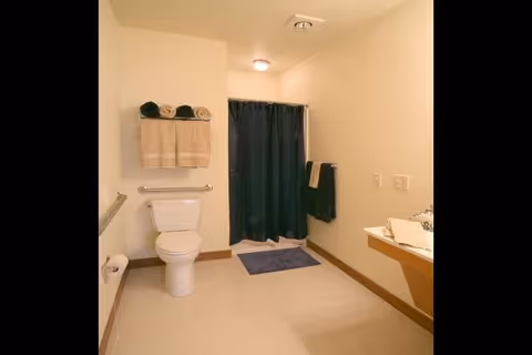 A clean, spacious bathroom with a white toilet, beige walls, and a shower area with a dark blue curtain. There are beige and dark blue towels hanging on racks, a blue bath mat on the floor, and a sink with a countertop on the right side.