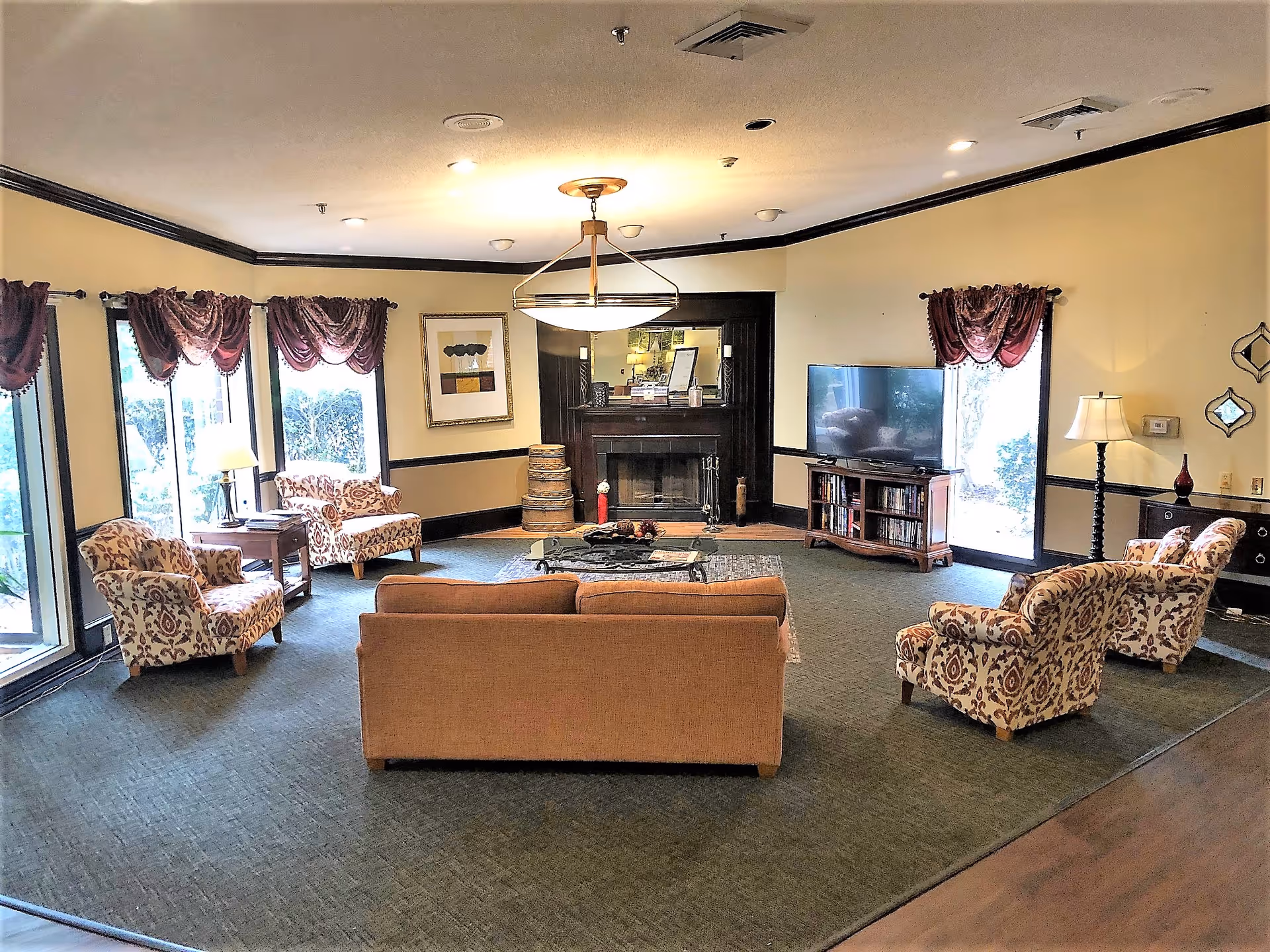 A spacious senior living community lounge with a beige sofa and four patterned armchairs arranged around a coffee table. The room features large windows with burgundy curtains, a fireplace with a mirror above it, a flat-screen TV on a wooden stand, and several lamps providing warm lighting.