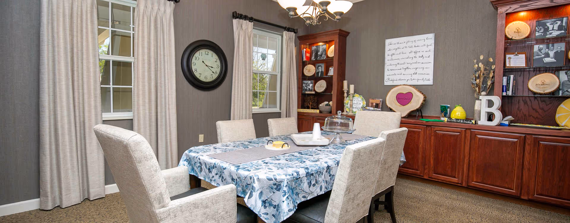 A dining room with a rectangular table covered with a blue and white floral tablecloth surrounded by six beige upholstered chairs. The room has two windows with beige curtains, a wall clock between the windows, and wooden built-in cabinets and shelves displaying decorative plates and items. A chandelier with five lights hangs above the table.