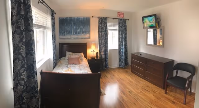 A small, neatly arranged bedroom with a single bed covered in a patterned quilt, a bedside table with a lamp, two windows with floral curtains, a dresser with a mounted TV above it, a chair, and a painting on the wall. The floor is wooden, and there is an exit sign above one window.