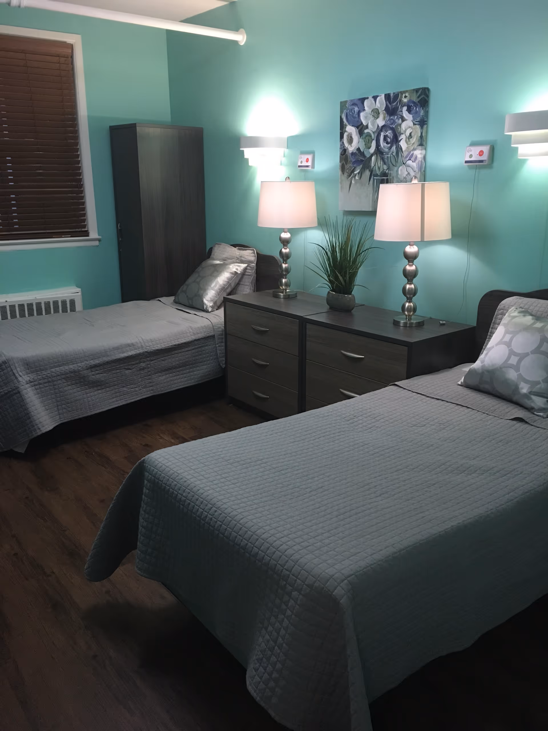 A neatly arranged twin bedroom with teal walls, two single beds, a dresser topped with lamps and a plant, and floral wall art.