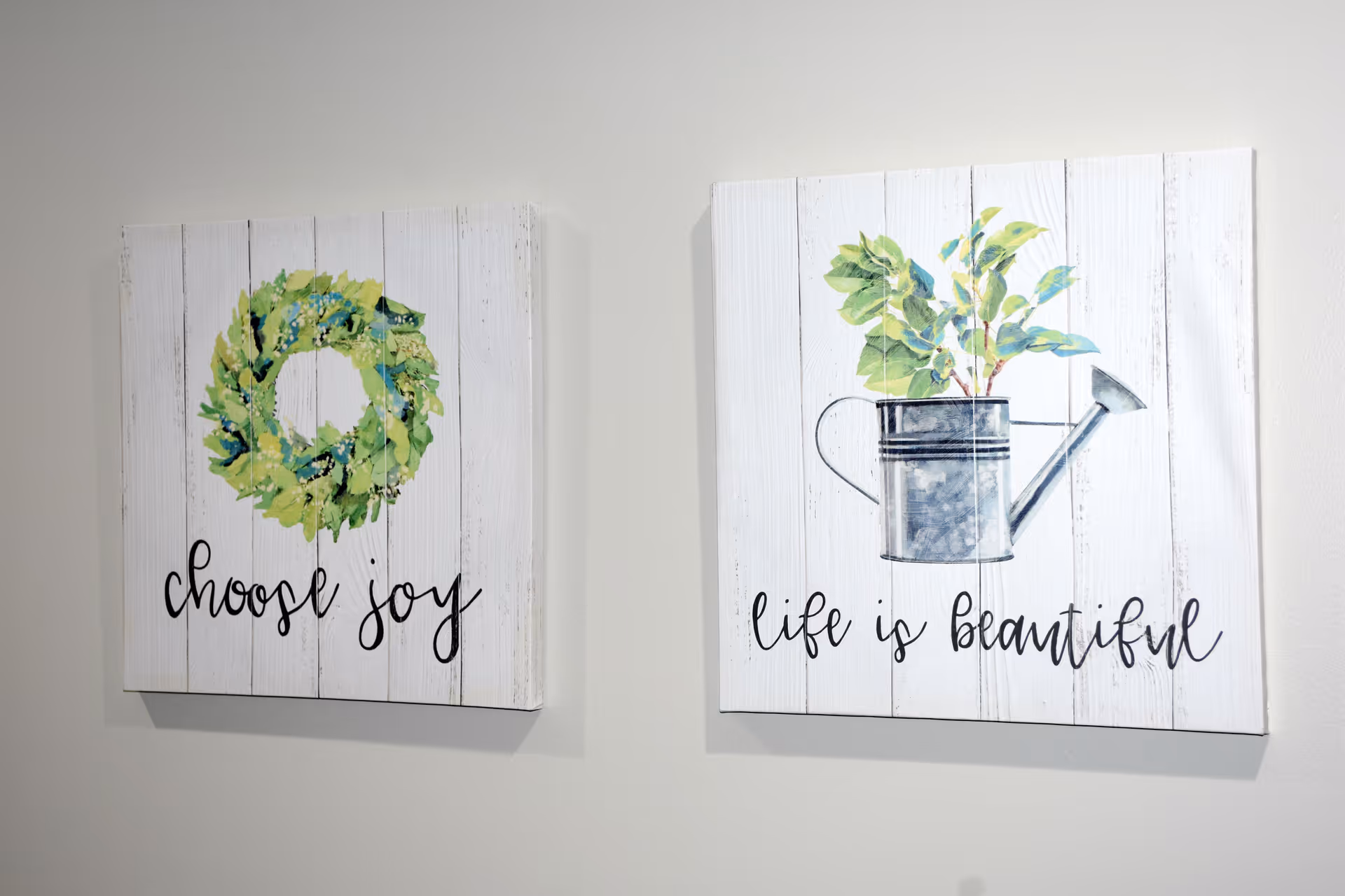 Two decorative wall art pieces on a white wall. The left piece features a green wreath with the words 'choose joy' written below it. The right piece shows a watering can with green leaves growing out of it and the words 'life is beautiful' written below.