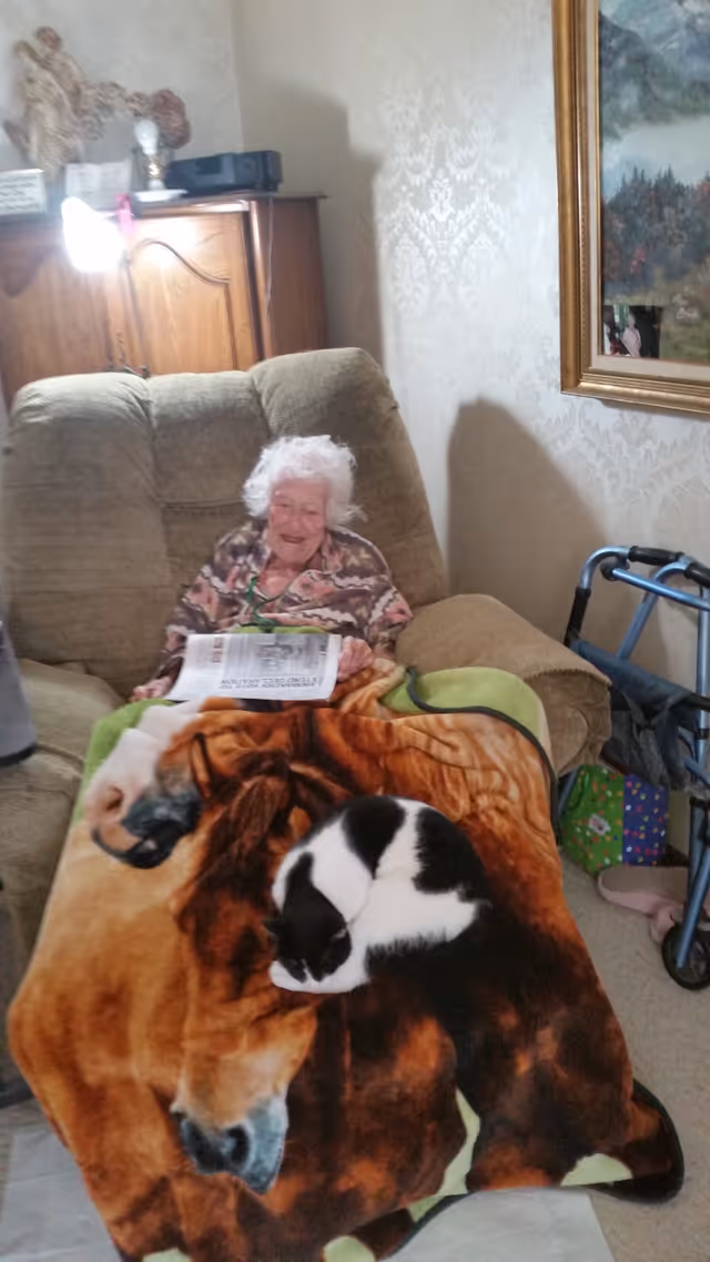 An elderly woman sitting in a recliner chair covered with a blanket featuring a horse design. A black and white cat is curled up sleeping on the blanket. The woman is holding a newspaper and smiling. There is a walker beside the chair, a wooden cabinet with decorative items, and a framed painting on the wall.