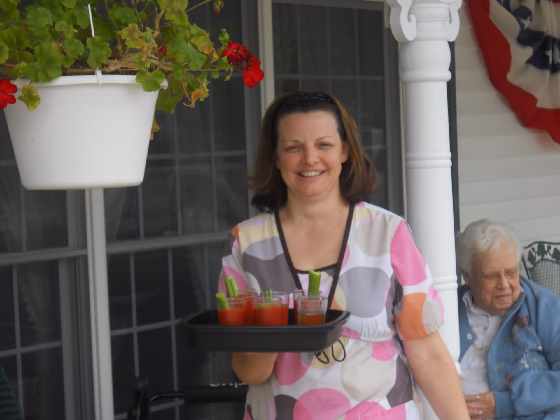 A smiling woman holding a tray with several glasses of red drinks garnished with celery stalks, standing outside near a white column and a hanging flower pot with red flowers. An elderly woman is seated in the background on a porch with white siding and a patriotic bunting decoration.