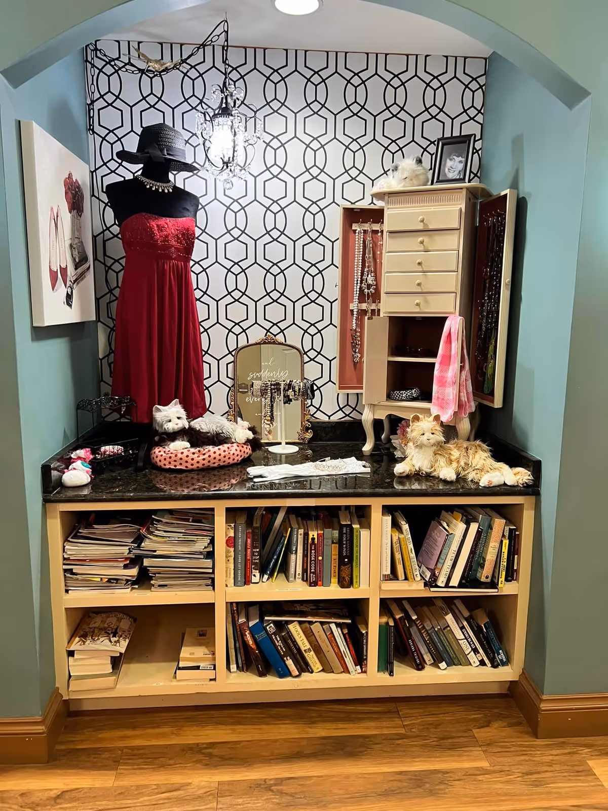 A decorative nook with a black mannequin dressed in a red strapless dress and black hat, a small chandelier hanging above, a jewelry cabinet with open doors displaying necklaces, several plush cat toys, a framed photo, and a bookshelf filled with books and magazines below. The back wall has a geometric black and white pattern, and the surrounding walls are painted teal.