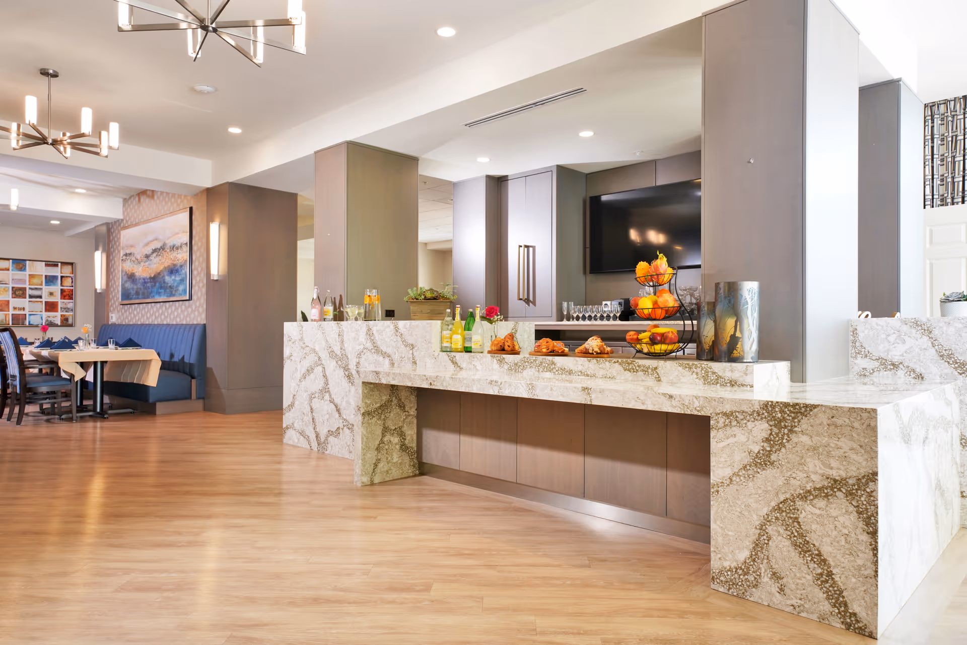 A spacious and modern dining area in Emerald Care Home featuring a large marble countertop with various beverages and pastries displayed. In the background, there are dining tables with blue cushioned seating, contemporary light fixtures, and decorative artwork on the walls.