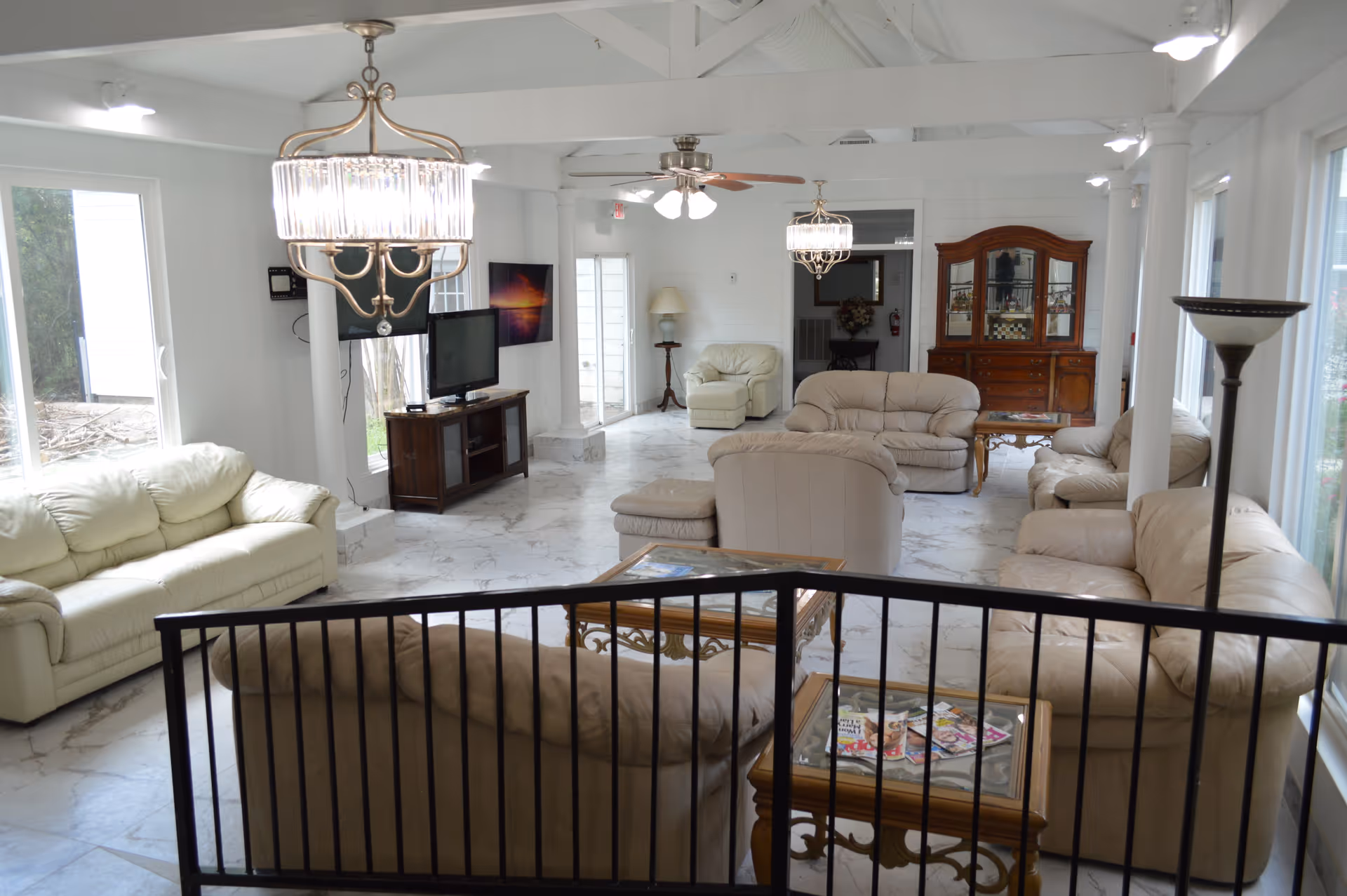 A spacious living room in an assisted living facility with multiple beige leather sofas and armchairs arranged around glass-top wooden coffee tables. The room has white walls, large windows letting in natural light, a ceiling fan, two chandeliers, a wooden cabinet with glass doors, a TV on a wooden stand, and a floor lamp. The floor is covered with white marble tiles.