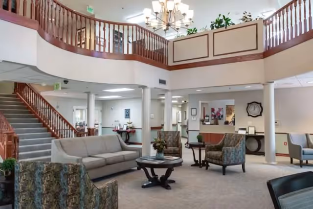 A bright, spacious senior living facility lobby with a curved second-floor balcony, staircase, chandeliers, and seating around a central coffee table.