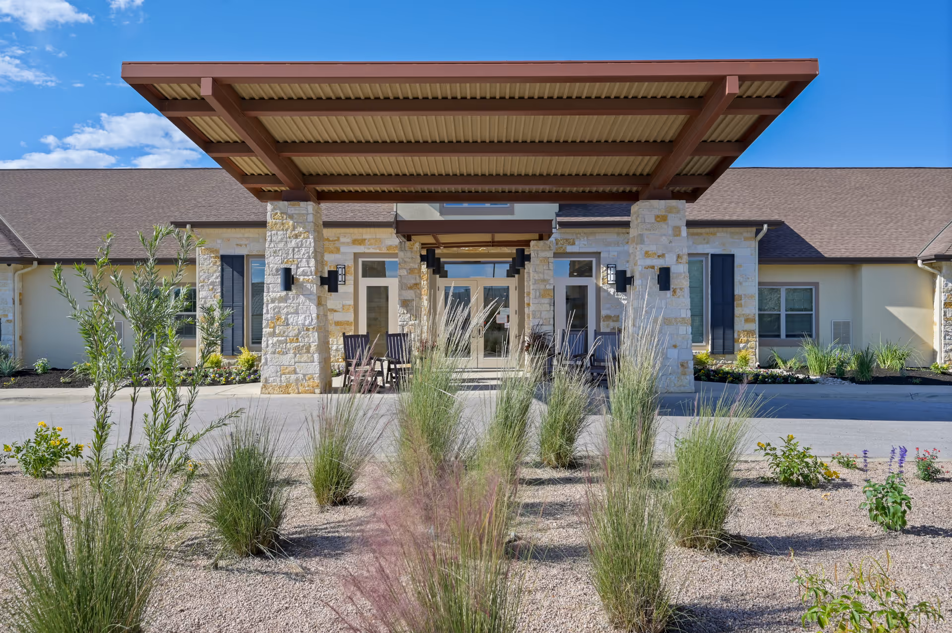 Front exterior view of The Monarch at Cedar Park building with a covered entrance supported by stone pillars, surrounded by landscaped plants and shrubs under a clear blue sky.