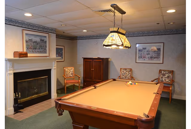 A pool table in a cozy communal game room with a fireplace, chairs, framed artwork, and a stained-glass pendant light.