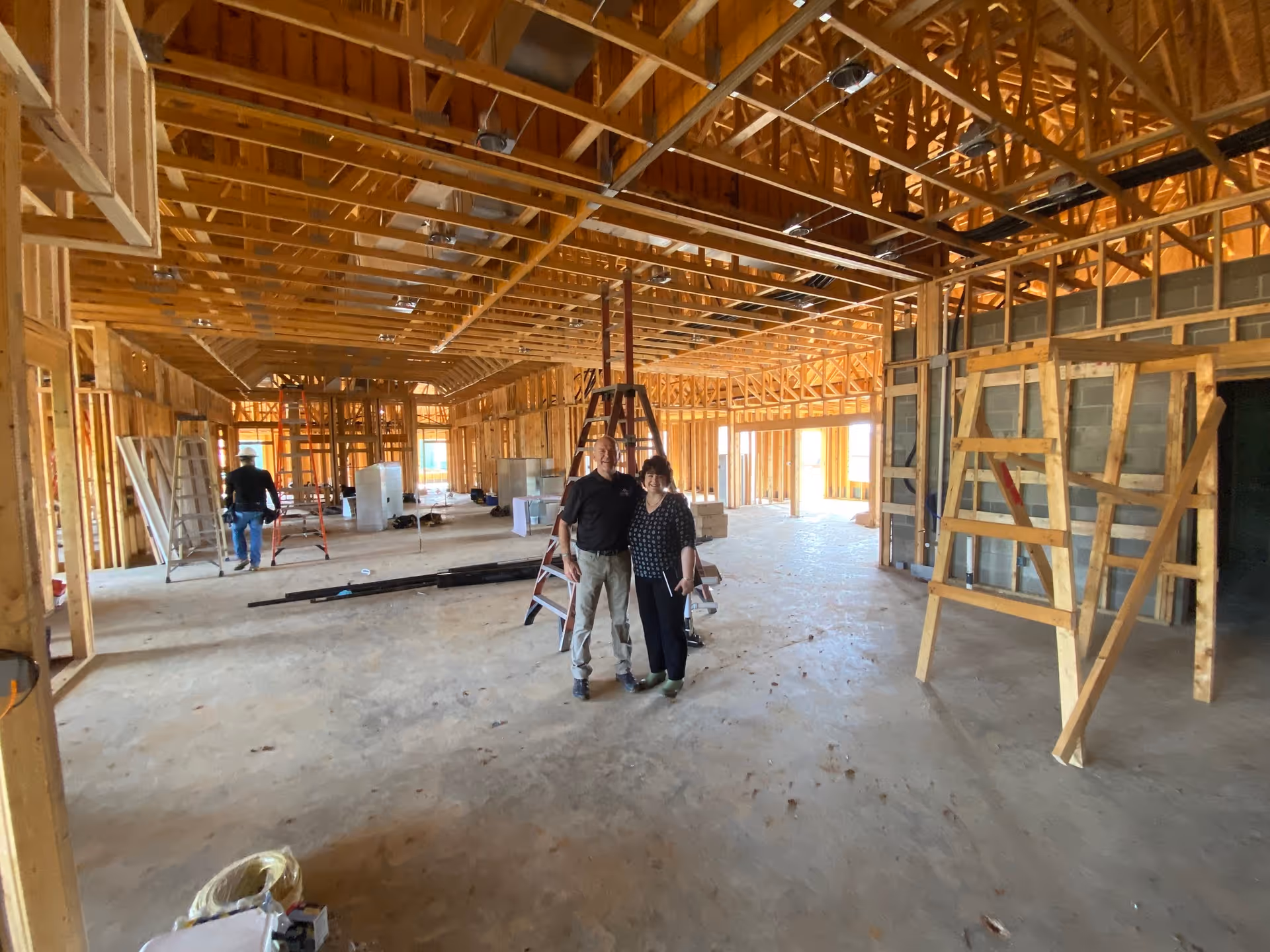 Interior view of a building under construction with exposed wooden framing and ceiling beams. Two people stand in the center near a ladder, and another person is seen walking away in the background. Construction materials and tools are scattered around the unfinished floor.