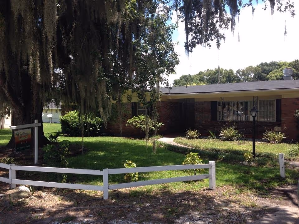 Exterior view of a single-story brick building with a black roof, surrounded by green grass, bushes, and a large tree with hanging moss. A white wooden fence runs along the front, and a sign near the tree reads 'Heritage ALF of Plant City'.