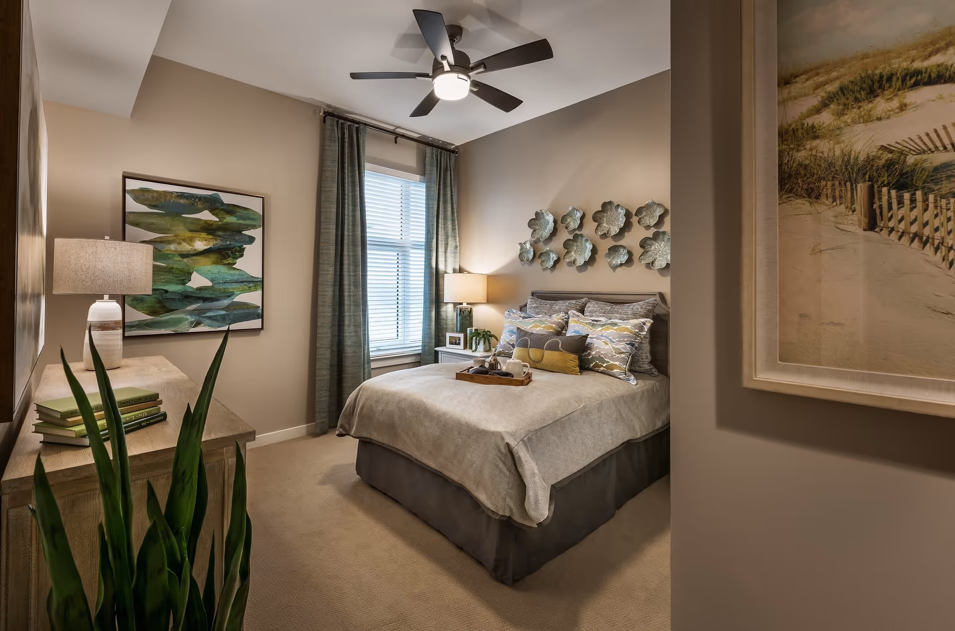A cozy bedroom with a neatly made bed featuring multiple decorative pillows and a tray on top. The room has beige walls, a ceiling fan with light, a window with green curtains, a wooden dresser with a lamp and books, and decorative wall art including metal flower shapes above the bed and a framed beach scene on the right wall.