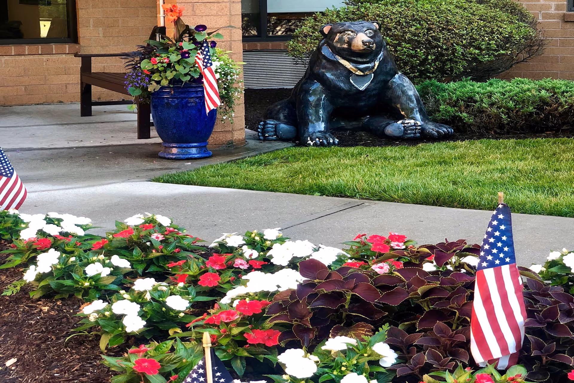 Outdoor garden area with a flower bed of red, white, and pink flowers, small American flags placed among the flowers, a large blue planter with colorful flowers and an American flag, a concrete walkway, and a black bear statue sitting on the grass near a brick building wall.