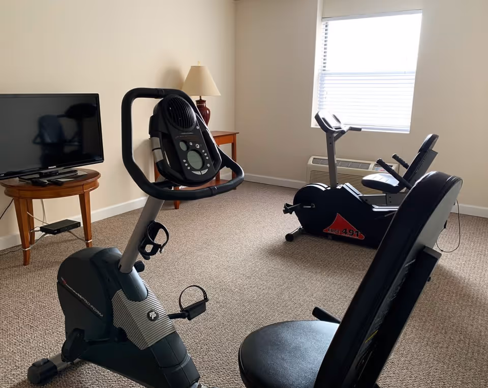 A small exercise room with two stationary exercise bikes facing a window with blinds. There is a TV on a wooden side table and a lamp on another small table against the wall. The room has beige walls and carpeted floor.