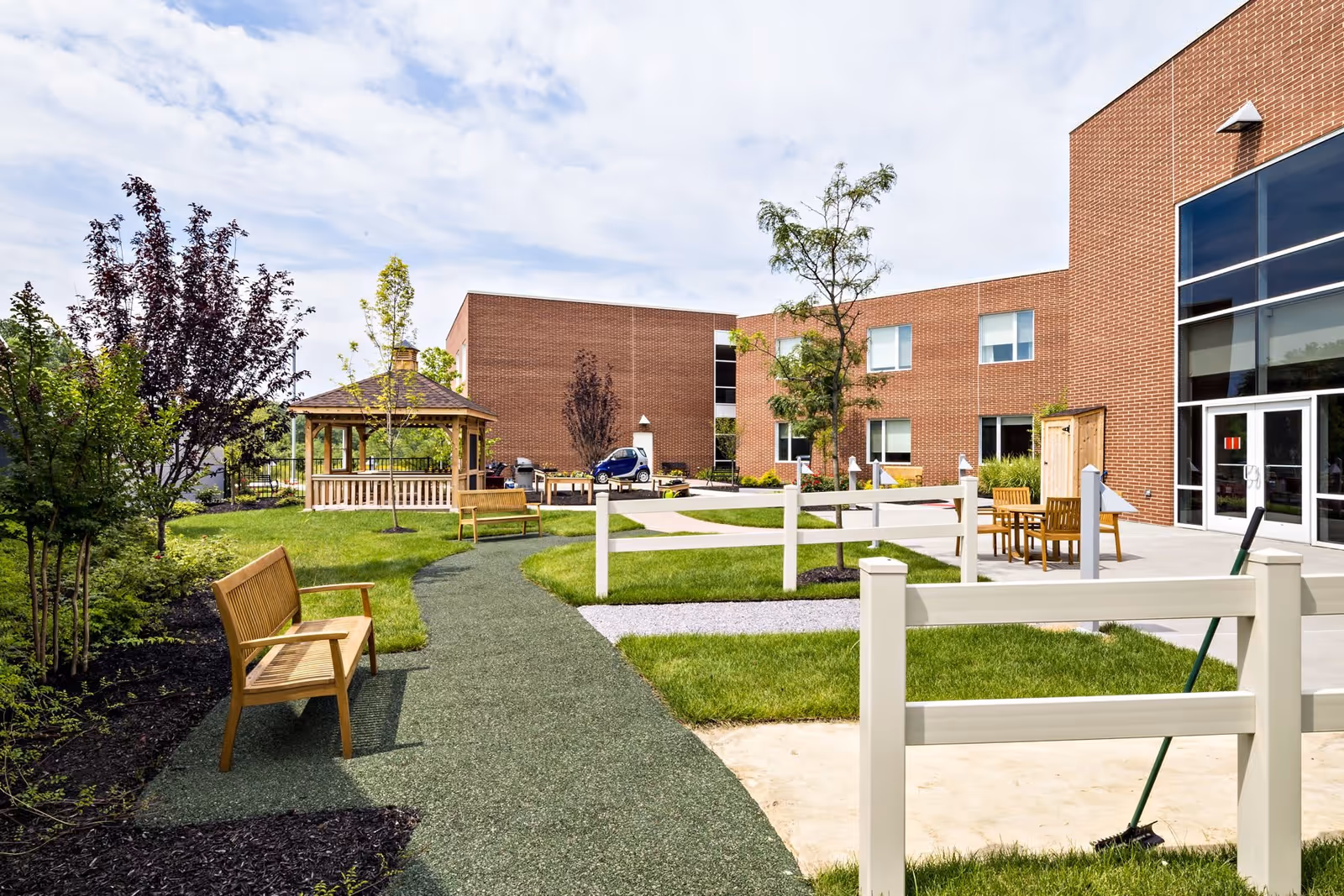 Outdoor courtyard area of Merwick Care & Rehabilitation Center featuring a paved walking path, wooden benches, a gazebo, trees, and a brick building in the background under a partly cloudy sky.