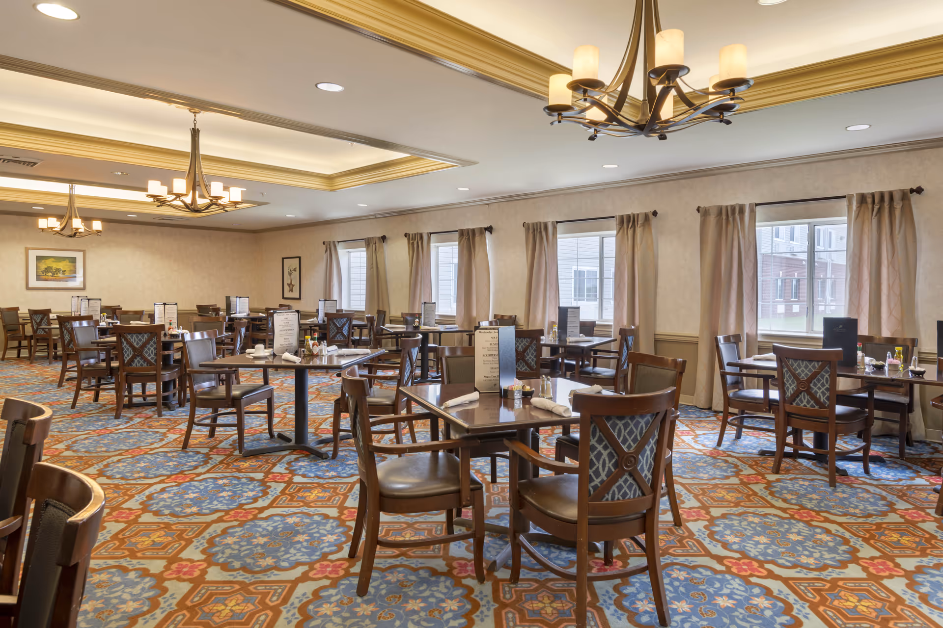 A spacious dining room with multiple wooden tables and chairs arranged neatly. The room features large windows with beige curtains, patterned carpet with blue and orange designs, and elegant chandeliers hanging from the ceiling. Menus and condiments are placed on the tables.