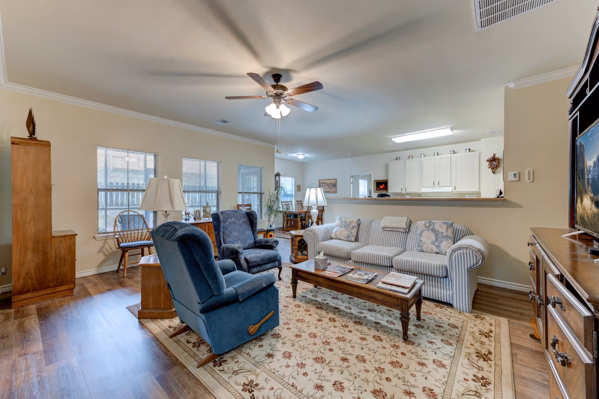 A cozy living room with a striped sofa, two blue armchairs, a wooden coffee table with magazines, a patterned area rug, and a ceiling fan with lights. The room has wooden flooring and large windows letting in natural light. In the background, there is a dining area and a kitchen with white cabinets.