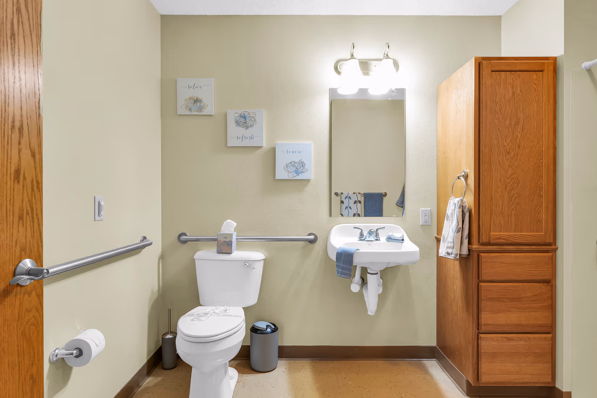 A clean and accessible bathroom with beige walls, a white toilet with grab bars on both sides, a white wall-mounted sink with a blue hand towel, a mirror with two lights above it, a wooden cabinet with drawers and a towel ring holding a patterned towel, and three decorative wall art pieces with floral designs and words 'relax', 'refresh', and 'renew'.