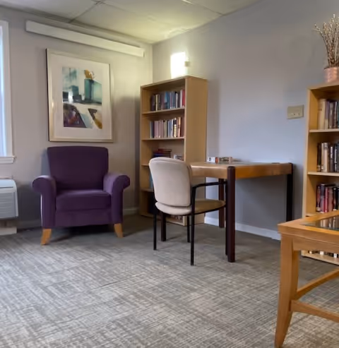 A cozy room with a purple armchair, a wooden table with a beige chair, two wooden bookshelves filled with books, a framed abstract painting on the wall, and a window with white trim. The room has a carpeted floor and soft lighting.