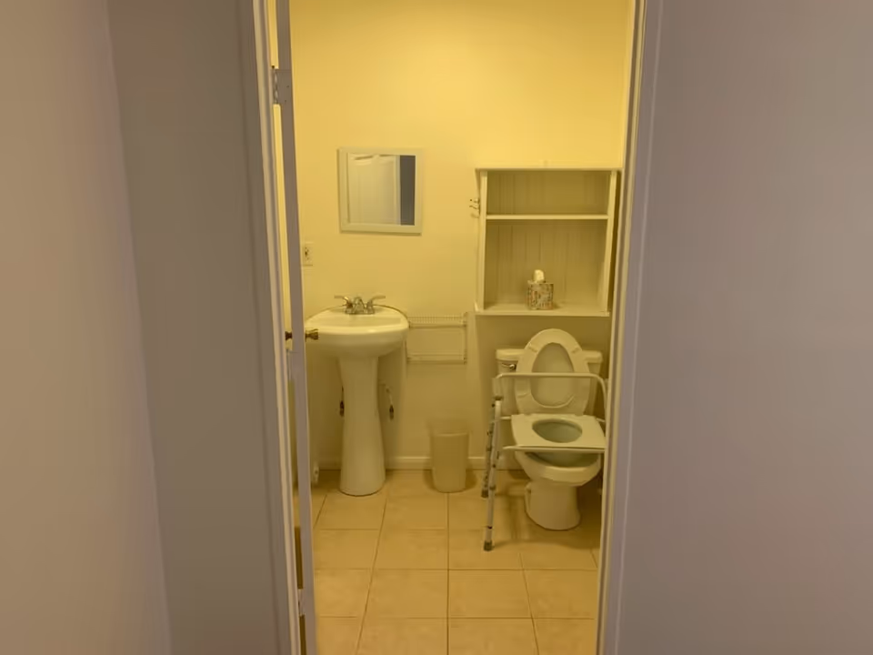 View through a doorway into a small bathroom with a pedestal sink, a mirror above it, a toilet with a raised seat and safety rails, a small shelf above the toilet holding a tissue box, and a waste bin on the tiled floor.