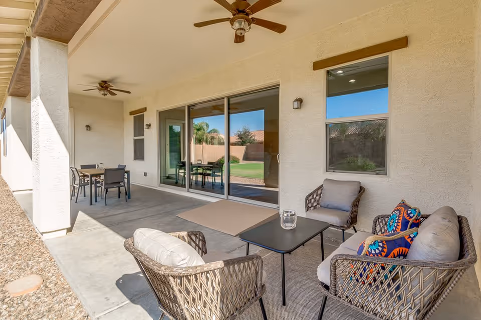 Covered outdoor patio area with wicker seating including a loveseat and two chairs with cushions and colorful pillows, a black coffee table, and a dining table with four chairs. Ceiling fans are mounted on the patio ceiling, and sliding glass doors lead inside to a room with a view of a green lawn and a fenced backyard.