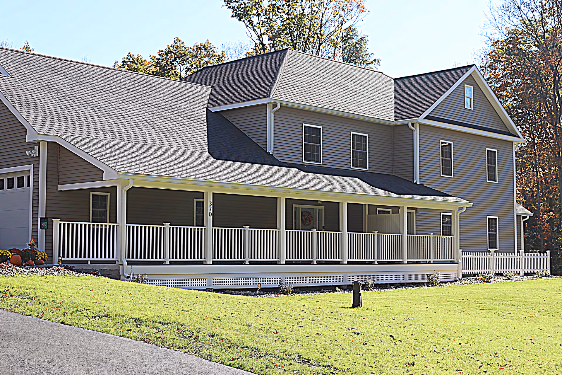 Exterior view of a two-story residential care home with gray siding, a large covered porch with white railings, and a well-maintained lawn in front. Trees with autumn foliage are visible in the background.