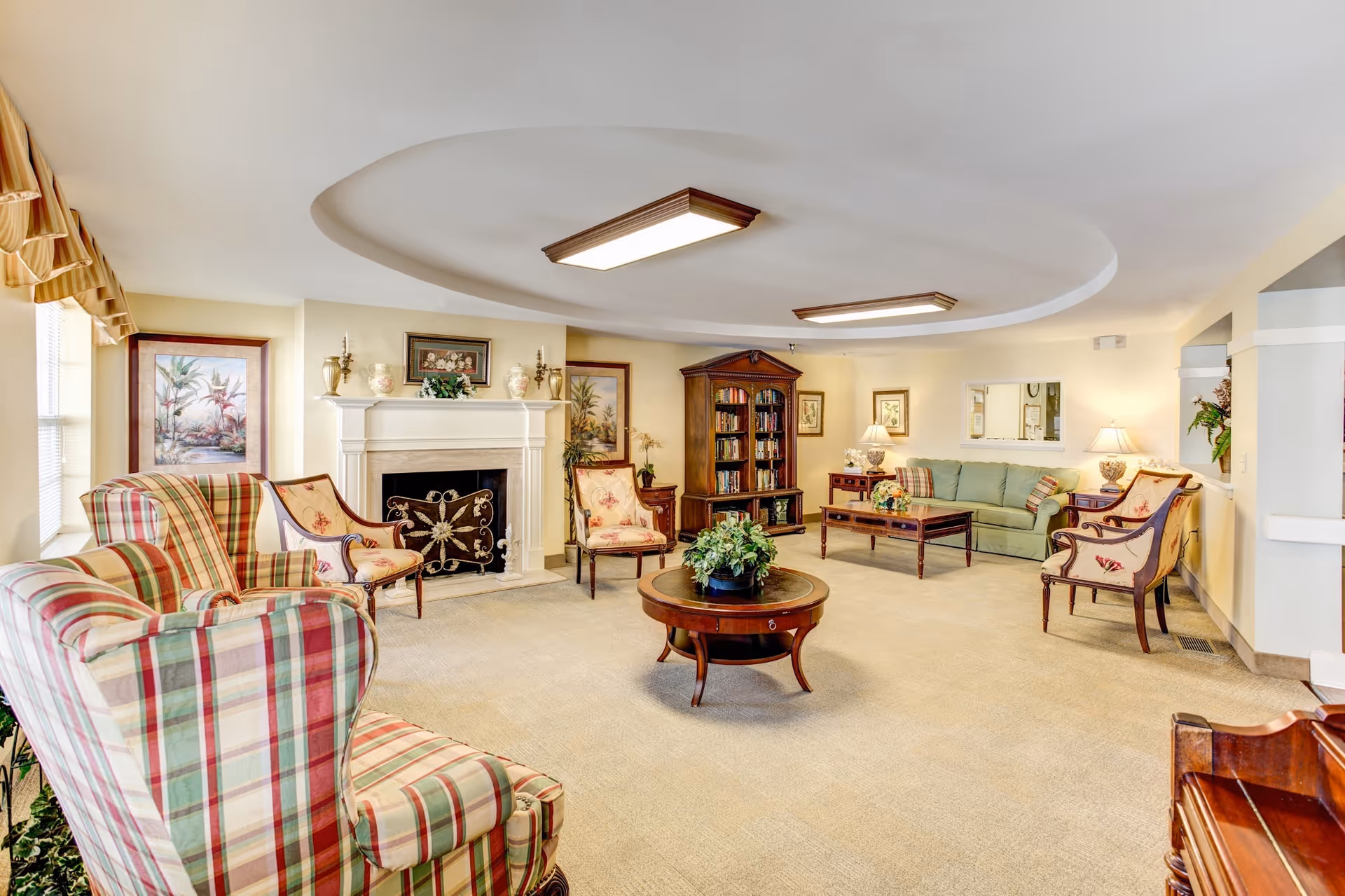 Bright communal living room with plaid armchairs, a sofa, coffee tables, a fireplace, and a bookshelf.